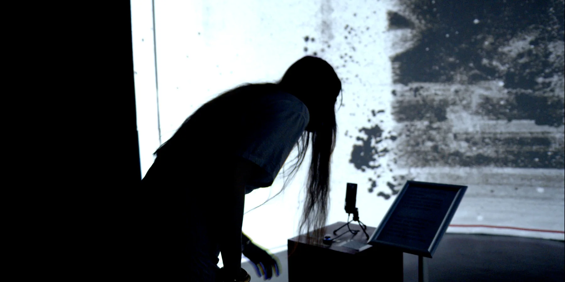 A long-haired visitor leans toward the microphone beside the projection